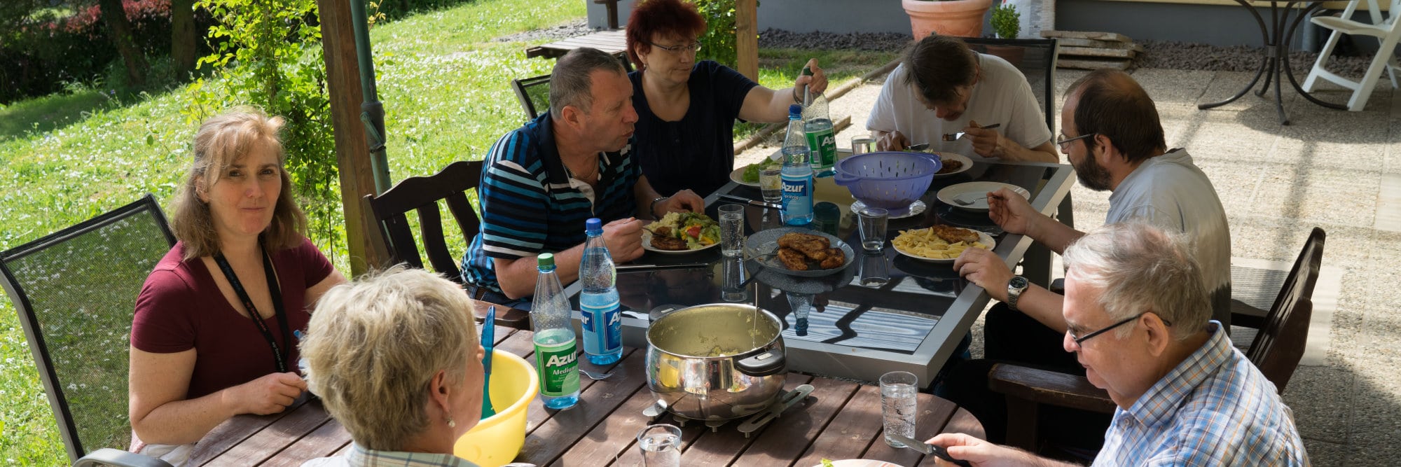 Eine Gruppe von Menschen sitzt auf der Terrasse an einem Tisch und nimmt gemeinsam eine Mahlzeit ein.