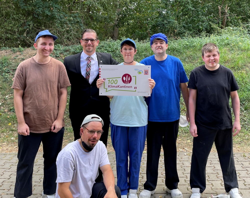 Vier Menschen, die in der Küche der bhw in den Wetterauer Werkstätten arbeiten, sind zu sehen. Der zweite Mann auf dem Foto ist Ulrich Buchwald, der die Küchen leitet. Die Frau in der Mitte hält ein Schild hoch. Auf dem Schild steht 100 Klima Kantinen. Die Menschen freuen sich über diese Auszeichnung.