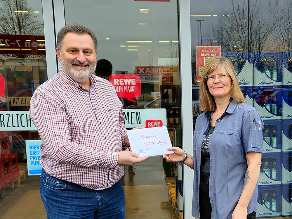 bhw-Geschäftsführer Markus Haselbauer steht links neben einer Frau. Sie halten zusammen ein Schild. Auf dem Schild steht: Spende 507,15 Euro. Beide stehen vorm Eingang vom REWE-Getränkemarkt.