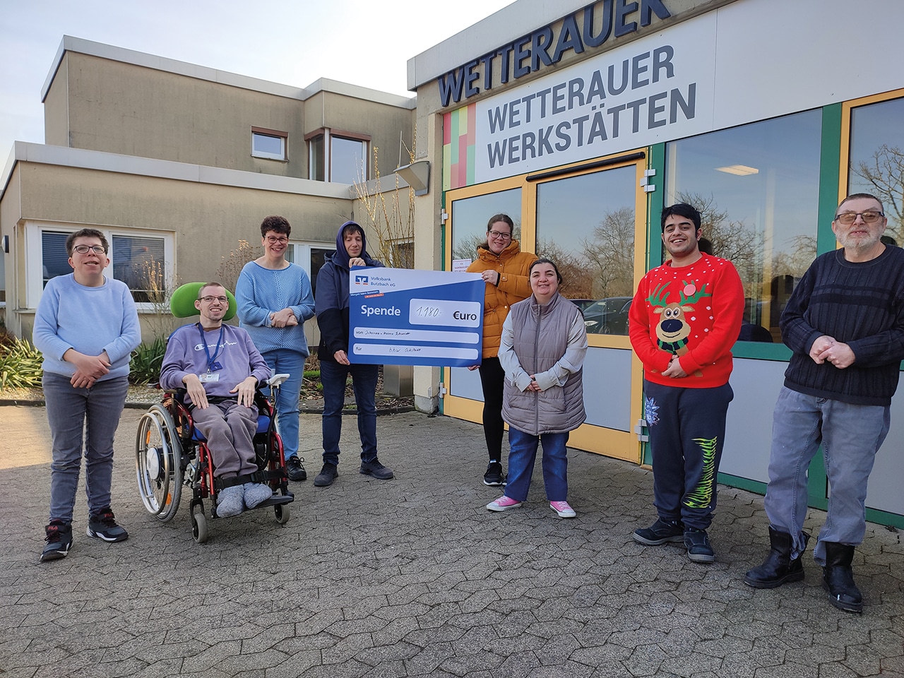 7 Personen stehen vor dem Eingang der Wetterauer Werkstätten. Ein Mann sitzt im Rollstuhl. Zwei Personen halten ein blaues Schild, auf dem steht: Spende 1180 Euro. Alle schauen in die Kamera und lachen.