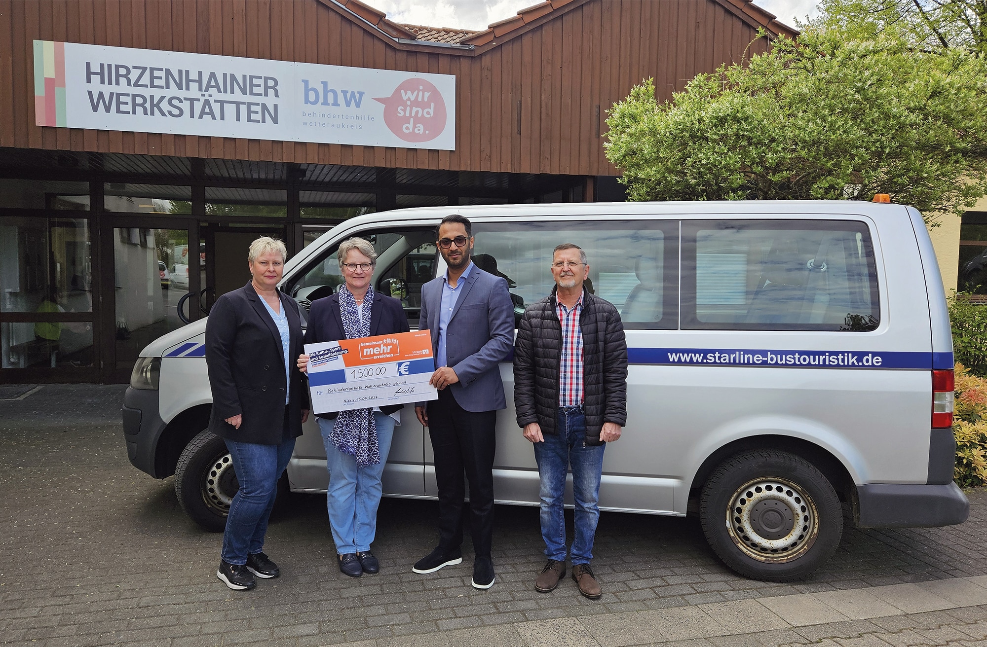 Vier Personen stehen vor einem silberfarbenen Bus. Die Frau und der Mann in der Mitte halten einen großen Spenden -Scheck. Im Hintergrund ist der Eingang von den Hirzenhainer Werkstätten zu sehen.